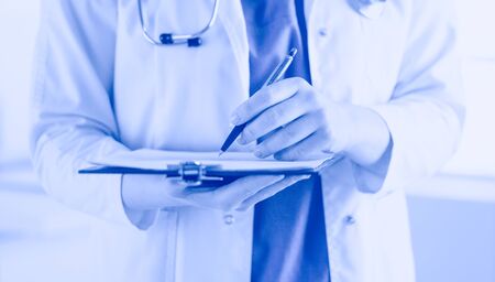 Doctor with a stethoscope, holding a notebook in his hand. Close-up of a female doctor filling up medical form at clipboard while standing straight in hospitalDoctor with a stethoscope, holding a notebook in his hand. Close-up of a female doctor filling up medical form at clipboard while standing straight in hospitalの写真素材