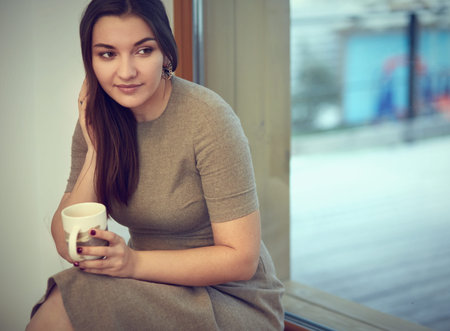 Young and cute lady sitting on the windowsill and looking out the window with cup of coffee