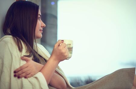 Young woman in plaid with cup of hot tea sitting on windowsillの写真素材
