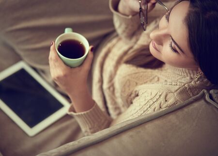 Happy young woman sitting on sofa holding a mug at home in the living roomの写真素材