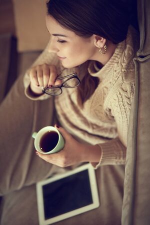 Happy young woman sitting on sofa holding a mug at home in the living roomの写真素材