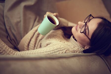 Young pretty woman having coffee and using laptop computer on sofa at homeの写真素材