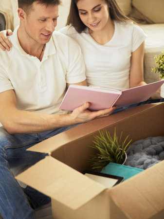 Cute couple unpacking cardboard boxes in their new home, sitting on the floor and looking at a...