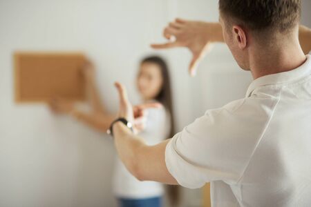 Woman helps her partner to hang painting on wall of their new house, she is framing the photo with her fingers.の写真素材
