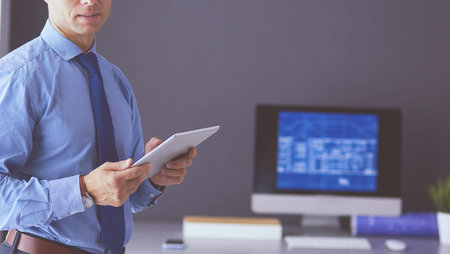 Smiling businessman with touchpad standing at workplace in officeの写真素材