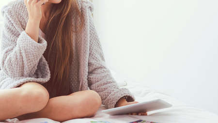 Relaxed young woman sitting on bed with a cup of coffee and digital tabletの写真素材