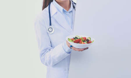 Portrait of a beautiful woman doctor holding a plate with fresh vegetables standing near blank. Woman doctorsの写真素材