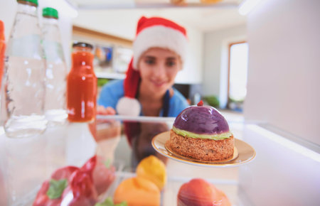 Portrait of female in santa hat standing near open fridge full of healthy food, vegetables and...