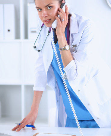 Young woman doctor in white coat at computer using phoneの写真素材