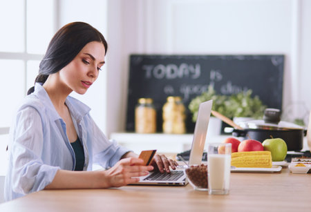 Smiling woman online shopping using computer and credit card in kitchenの写真素材