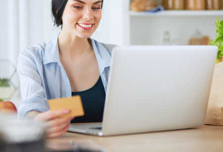 Smiling woman online shopping using computer and credit card in kitchenの写真素材
