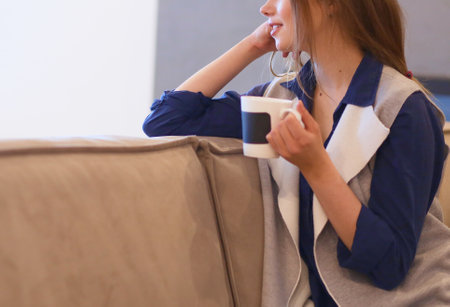 Young woman sitting on comfortable sofa with cup on coffee in hands in white roomの写真素材