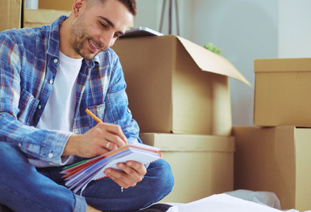 A moving man sitting on the floor in empty apartment, Among the Boxes, Checking the List of Thingsの写真素材