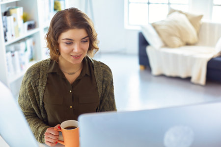 Attractive smiling woman sitting at office desk, holding a cup of coffee, she is relaxing and looking awayの写真素材