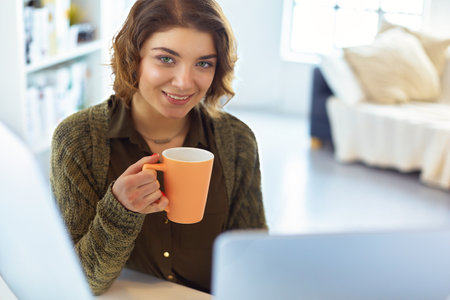Attractive smiling woman sitting at office desk, holding a cup of coffee, she is talking to someone by messenger.の写真素材