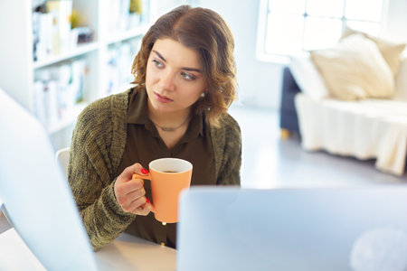 Attractive smiling woman sitting at office desk, holding a cup of coffee, she is relaxing and looking awayの写真素材