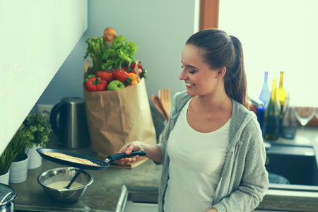 Young woman cooking pancakes at kitchen standing near stoveの写真素材