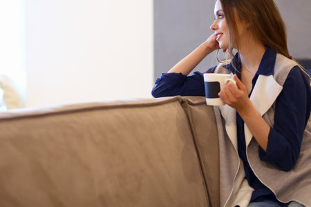 Young woman sitting on comfortable sofa with cup on coffee in hands in white roomの写真素材