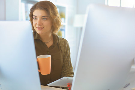 Attractive smiling woman sitting at office desk, holding a cup of coffee, she is relaxing and looking awayの写真素材