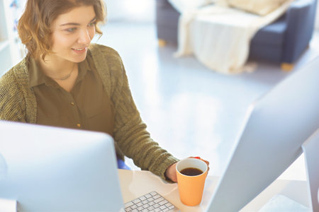 Attractive smiling woman sitting at office desk, holding a cup of coffee, she is relaxing and looking awayの写真素材