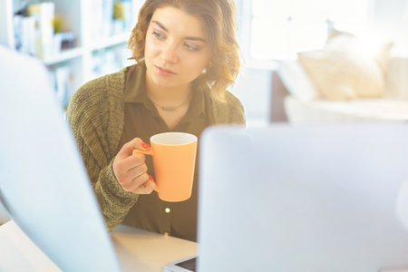 Attractive smiling woman sitting at office desk, holding a cup of coffee, she is relaxing and looking awayの写真素材