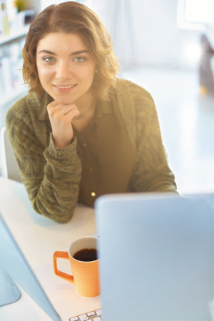 Attractive smiling woman sitting at office desk, holding a cup of coffee, she is relaxing and looking awayの写真素材