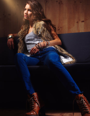 Portrait of elegant woman sitting on black sofa wearing a blue jeans and fur vestの写真素材