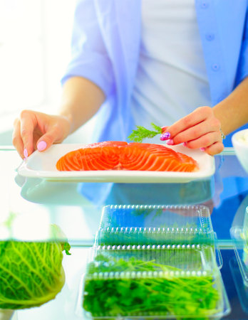 Portrait of female standing near open fridge full of healthy food, vegetables and fruits.の写真素材