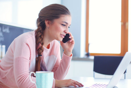Young woman sits at the kitchen table using a laptop and talking on a cell phone.の写真素材