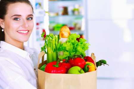 Young woman holding grocery shopping bag with vegetables .Standing in the kitchen. Woman in the kitchen looking at the cameraの写真素材