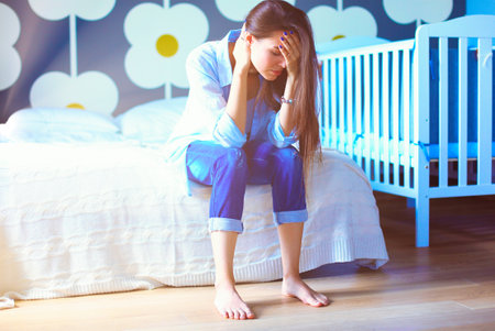 Young tired woman sitting on the bed near childrens cot. Young mom.の写真素材