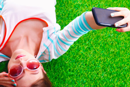 Beautiful young woman making selfie by her phone while lying in green grassの写真素材