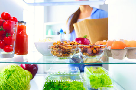 Portrait of female standing near open fridge full of healthy food, vegetables and fruitsの写真素材
