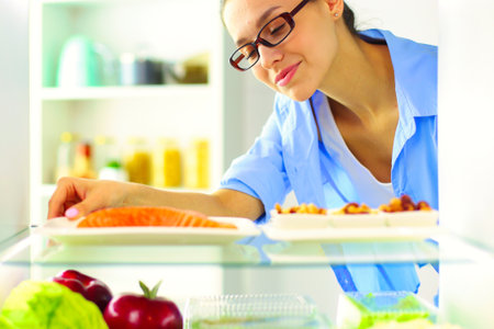 Portrait of female standing near open fridge full of healthy food, vegetables and fruitsの写真素材