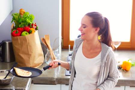 Young woman prepares pancakes in the kitchen while standing near the table. Woman in the kitchen. Cooking at kitchen.の写真素材