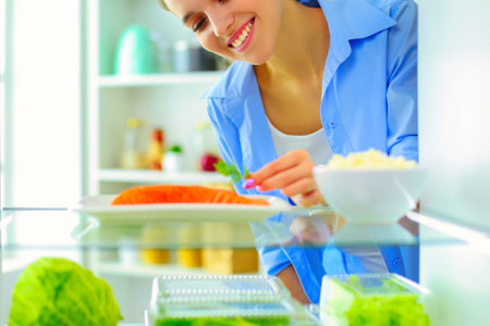 Portrait of female standing near open fridge full of healthy food, vegetables and fruitsの写真素材