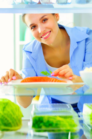 Portrait of female standing near open fridge full of healthy food, vegetables and fruitsの写真素材