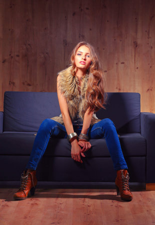 Portrait of elegant woman sitting on black sofa wearing a blue jeans and fur vestの写真素材