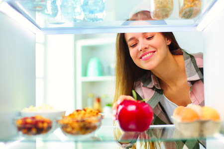 Portrait of female standing near open fridge full of healthy food, vegetables and fruitsの写真素材