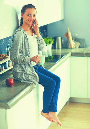 Woman using mobile phone sitting in modern kitchenの写真素材