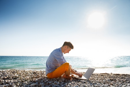 Handsome young barefooted man walking with laptop computer by the sea coastの写真素材