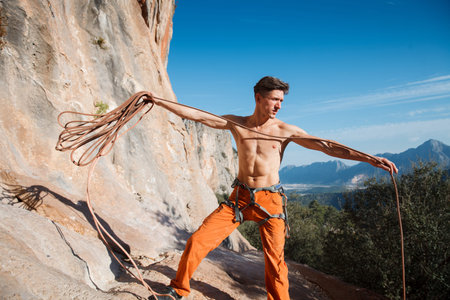 Handsome adult male rock climber collecting the belay rope over the blue sky and mountains. Turkey,...