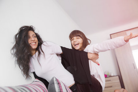 Young woman piggybacking her joyful little daughter at home in white lit room against the window....
