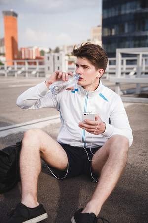 Adult male athlete drinking water against office buildings after urban running. Concept of hard working and active living, freelance, remote business, healthcare and wellbeingの写真素材