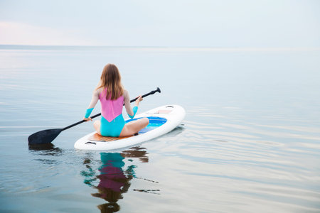 Unrecognizable young woman paddling on sup board with paddle. Sitting pose - concept of harmony with...