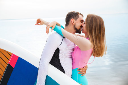 Young couple hugging and kissing on the beach with sup board. Side view closeup - concept of harmony with the nature, free and healthy living, freelance, remote business.の写真素材