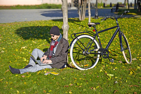 Young man working on a laptop in an autumn park.  His bicycle standing next to him.の写真素材