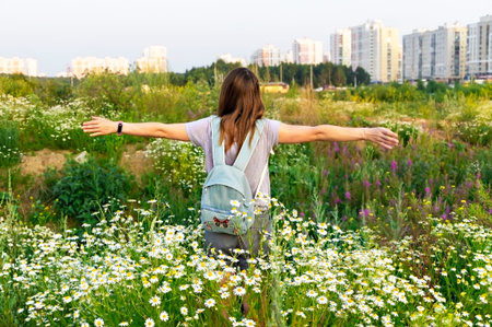Young caucasian woman with backpack standing from behind in a meadow among chamomile flowers with...