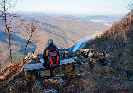 young man with a backpack from back sitting on a bench and looking at Drina river canyon, Serbia. Landscape, hike, tourism. Traveling aloneの写真素材