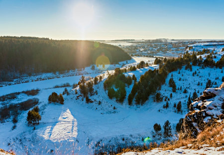 Winter landscape with a snow-covered river, cliffs with a pine forest and a village at golden hour...
